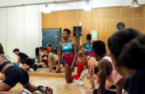 Une salle de danse avec un groupe de personnes en tenue de sport effectuant un étirement au sol. Au centre, une femme portant une brassière bleu métallique et un short rose est à genoux, semblant guider l'exercice. Un grand miroir sur le mur reflète les participants, qui sont divers en termes d'âge, de style et de couleur de peau. L'ambiance est dynamique et conviviale.